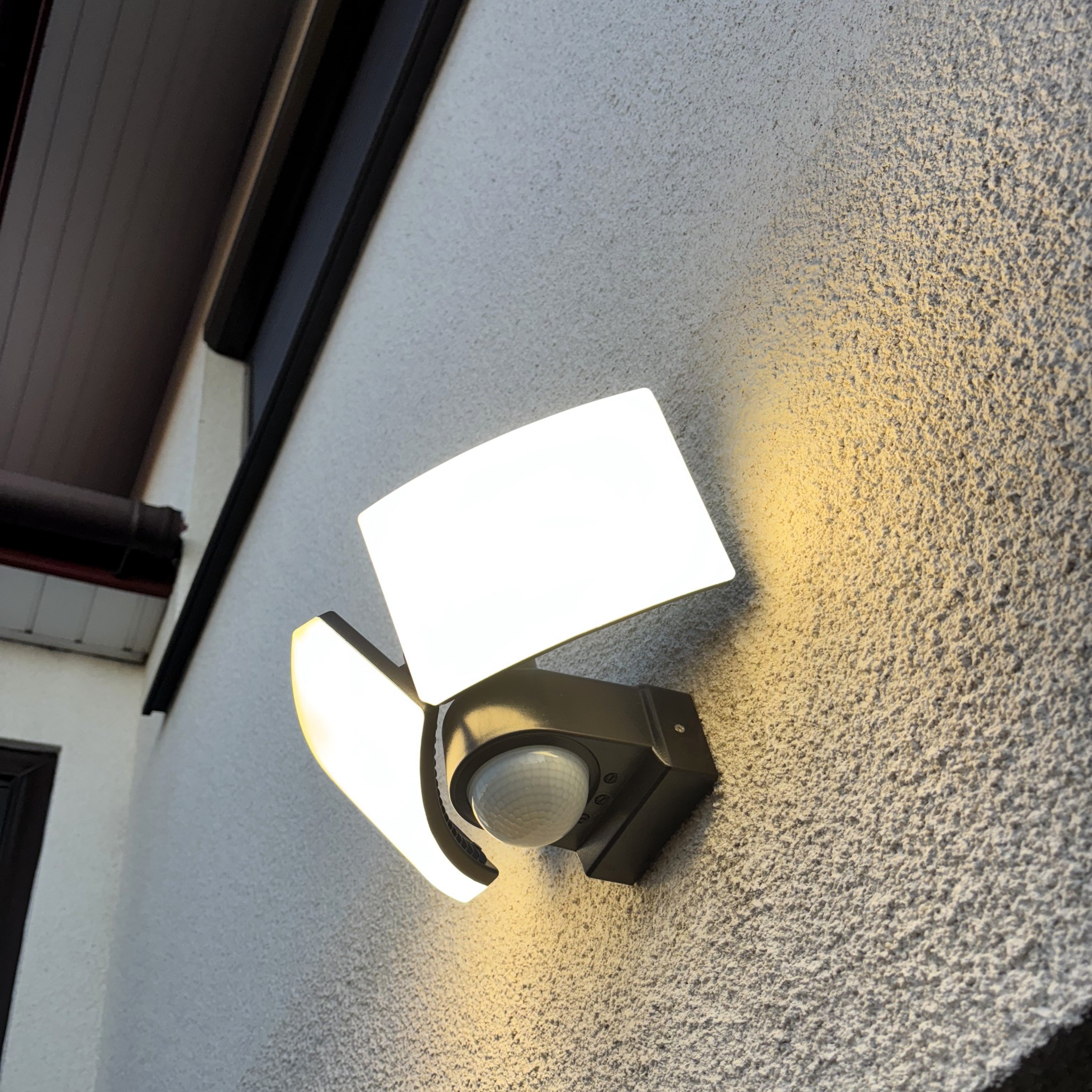 Zewnętrzna lampa LED z czujnikiem ruchu, zamontowana na teksturowanej ścianie budynku w Brzesku. Dwa regulowane panele świetlne emitują jasne, ciepłe światło.
