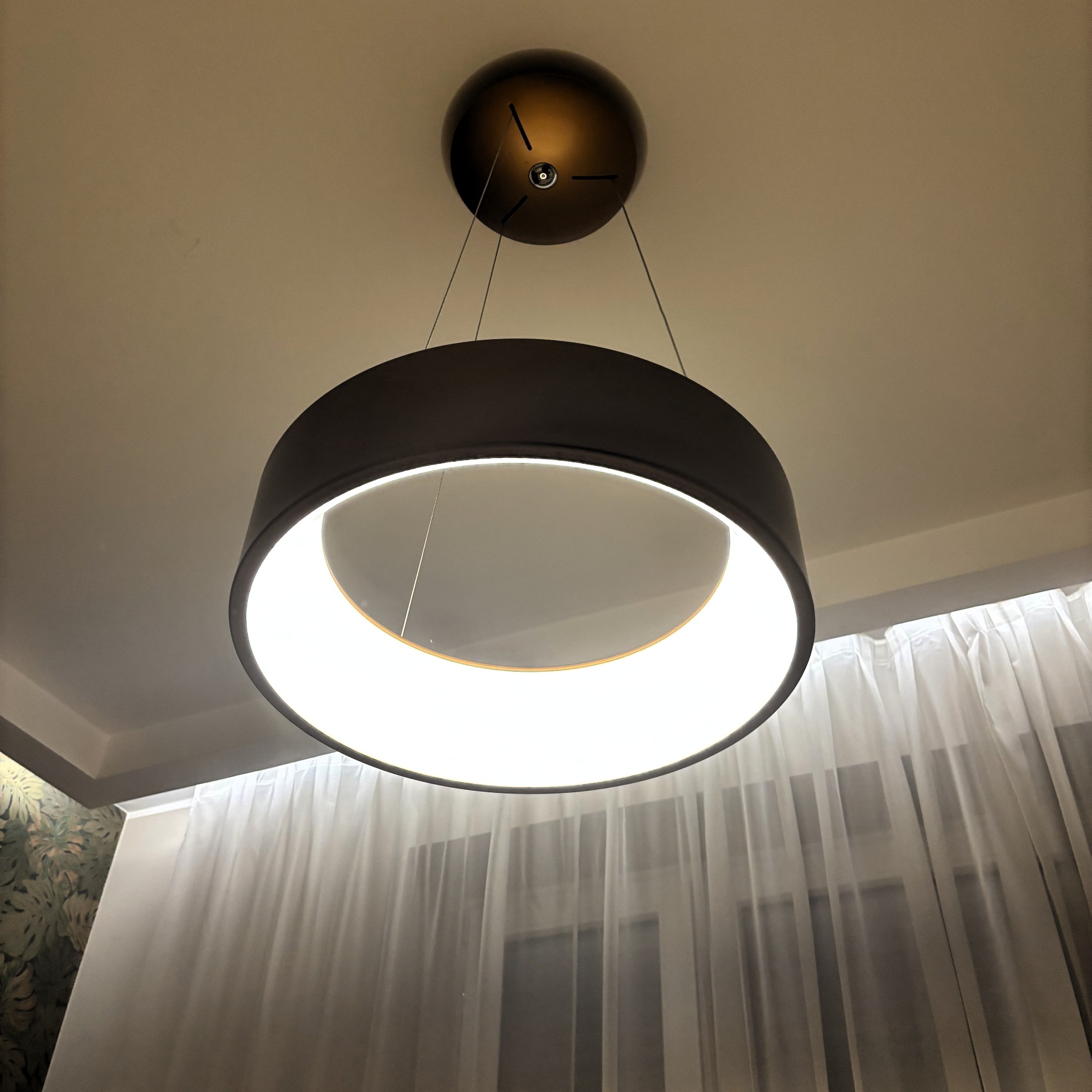 Nowoczesna, okrągła lampa sufitowa z wbudowanym oświetleniem LED, zawieszona na cienkich linkach, na tle okna z delikatnymi firankami.