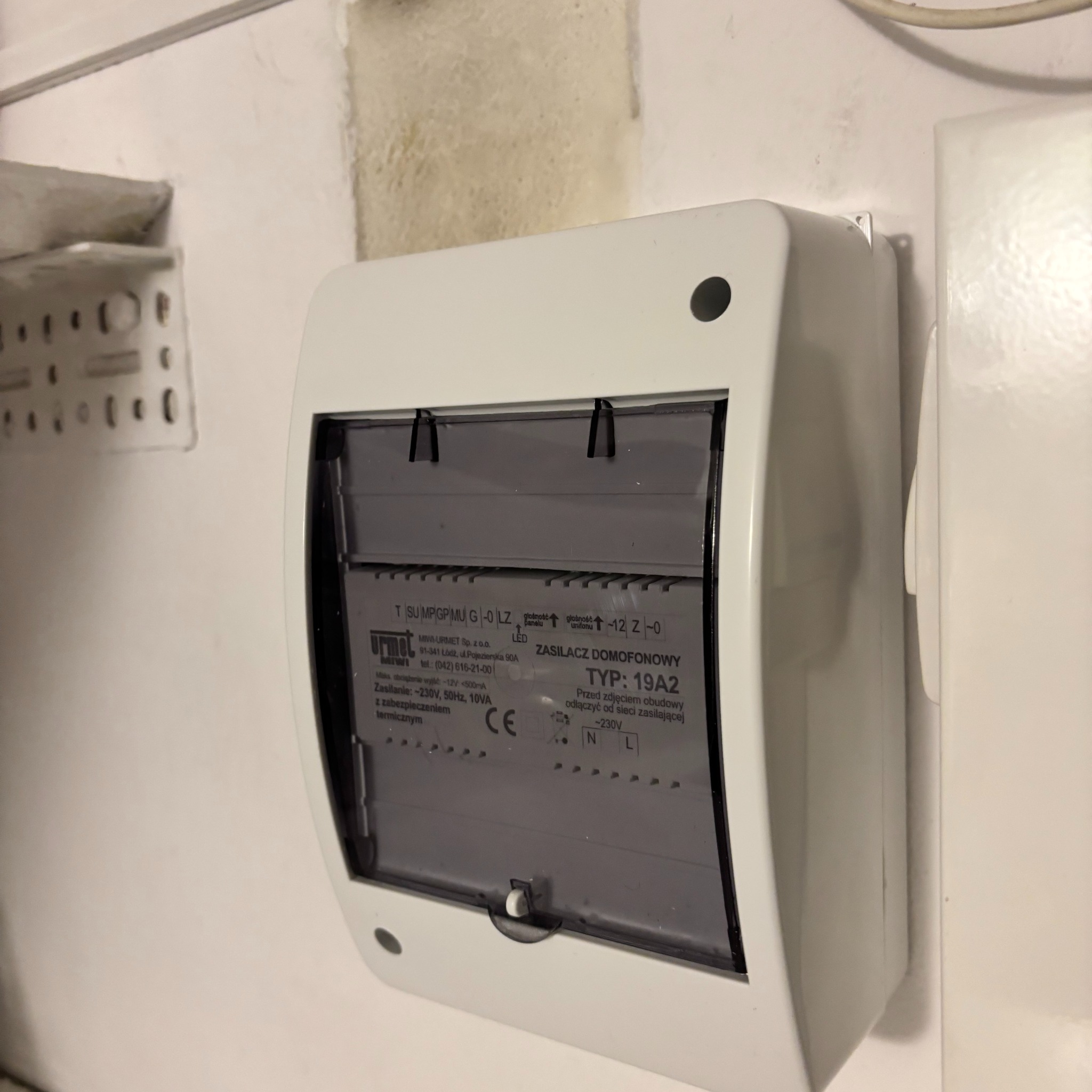 Zamknięty, biały zasilacz domofonowy Urmet 19A2, zamontowany na ścianie. Widoczne parametry techniczne i oznaczenia CE. Ujęcie z bliska, lekko z boku.