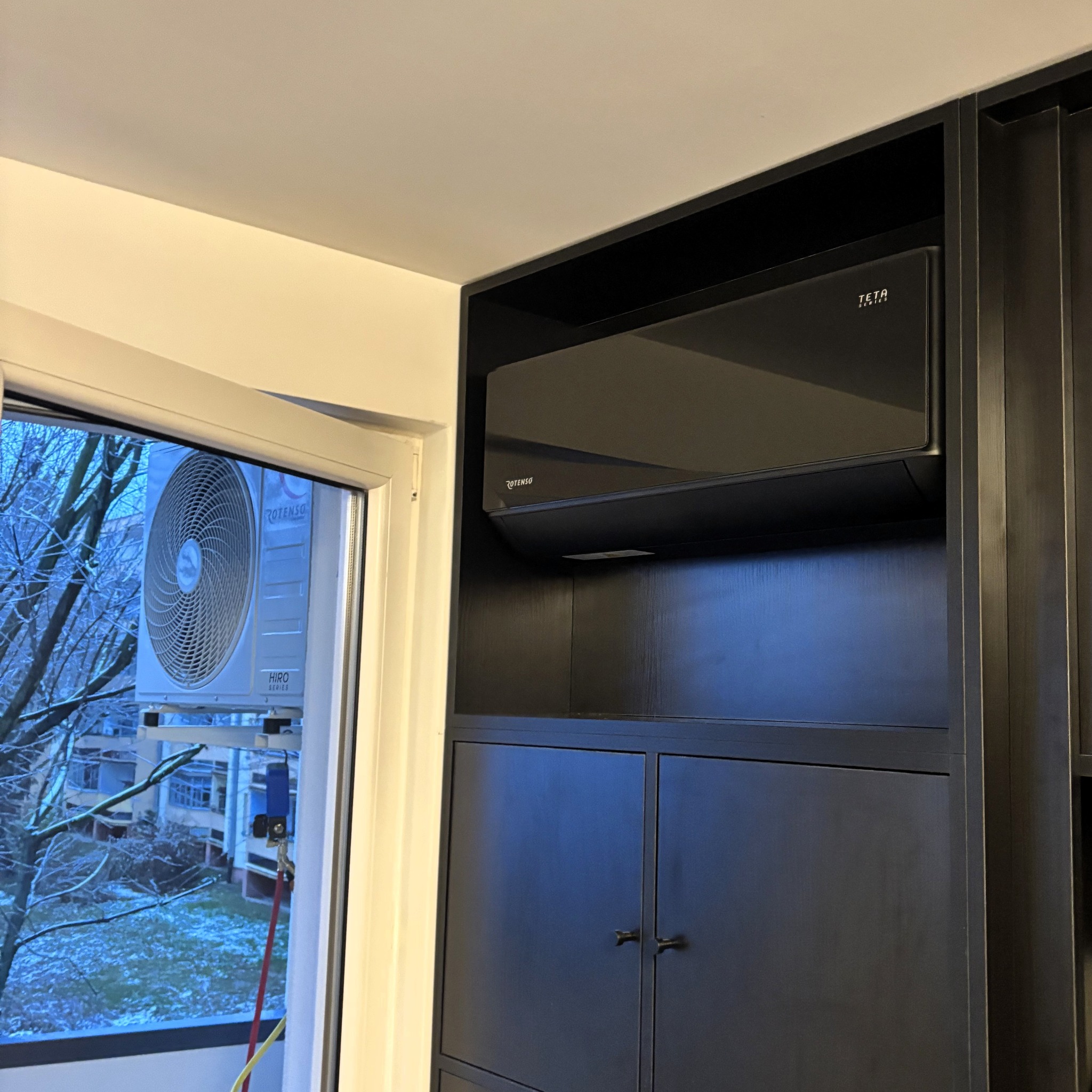 Czarny klimatyzator Rotenso Teta Series wkomponowany w ciemną zabudowę meblową, z widoczną jednostką zewnętrzną za oknem. Minimalistyczny design, nowoczesne rozwiązanie.