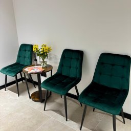 PsychoNovatis Centrum Terapii - Poczekalnia: trzy zielone krzesła z aksamitu, stolik z żółtymi kwiatami i magazynem. Stonowane kolory, minimalistyczny wystrój, płytki podłogowe.