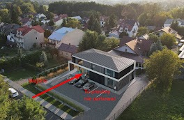 Nowoczesny, dwukondygnacyjny budynek z czarną elewacją i dużymi oknami, otoczony zielenią i zabudową jednorodzinną, widziany z lotu ptaka w słoneczny dzień.