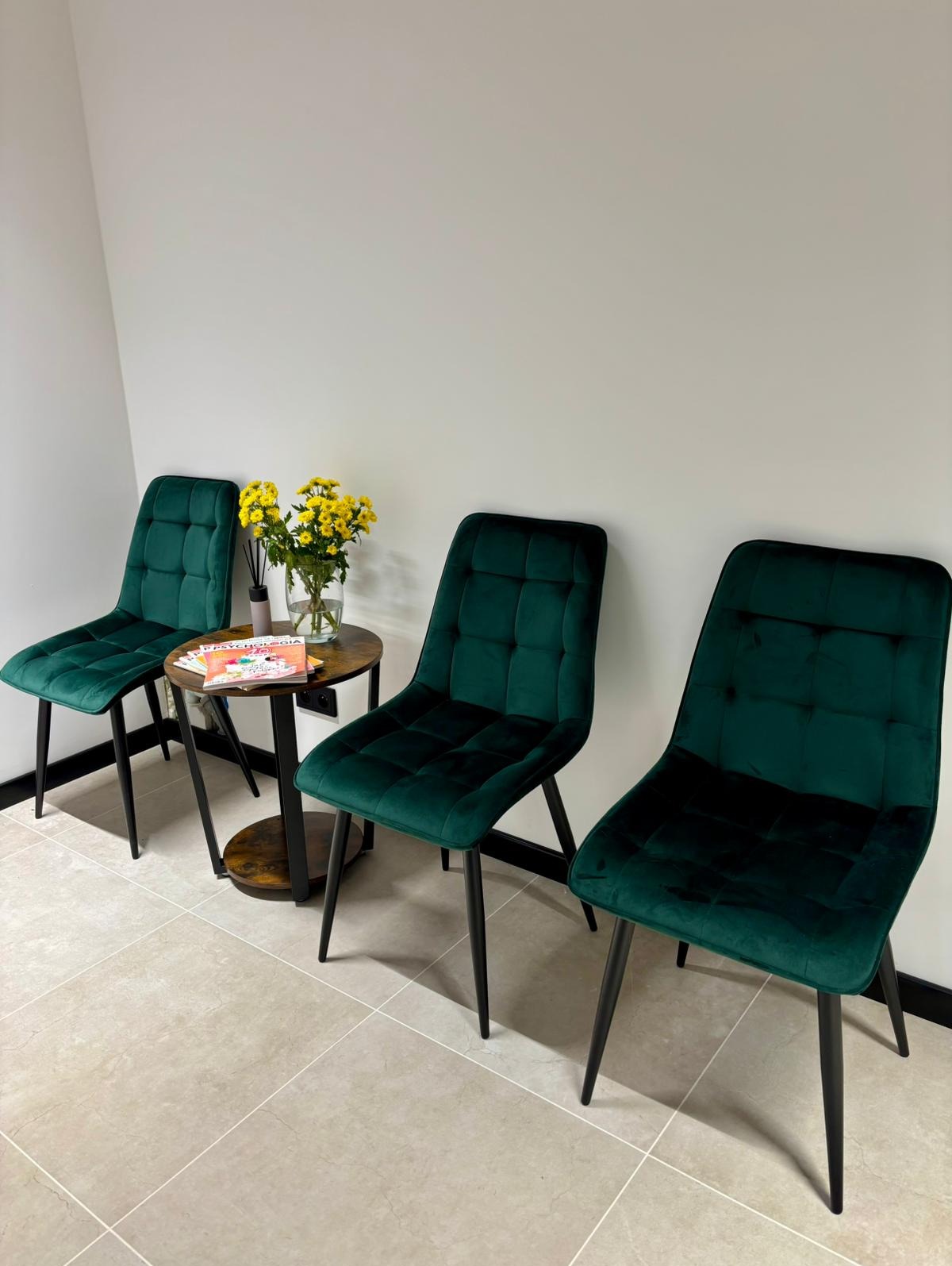 Poczekalnia: trzy zielone krzesła z aksamitu, stolik z żółtymi kwiatami i magazynem. Stonowane kolory, minimalistyczny wystrój, płytki podłogowe.