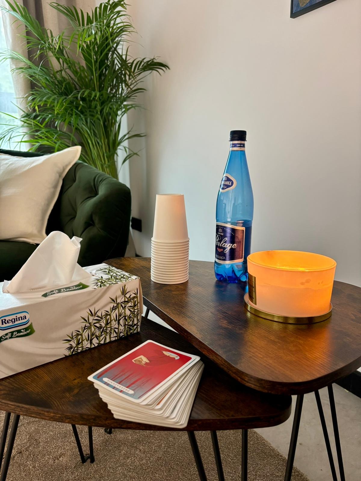 Gabinet terapeutyczny: karty, chusteczki, woda i świeca na stoliku kawowym, obok zielona sofa i roślina doniczkowa. Przytulna atmosfera.