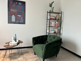 Jasne wnętrze gabinetu z zielonym fotelem i stolikiem. Na ścianie obraz w stylu abstrakcyjnym, obok regał z książkami i rośliną. Stonowana kolorystyka sprzyja relaksacji.