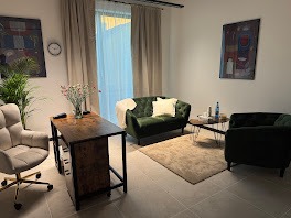 Jasny gabinet z zieloną sofą i fotelem, biurkiem z kwiatami, neutralnym dywanem i obrazami na ścianach. Przytulne wnętrze, gotowe na spotkanie.