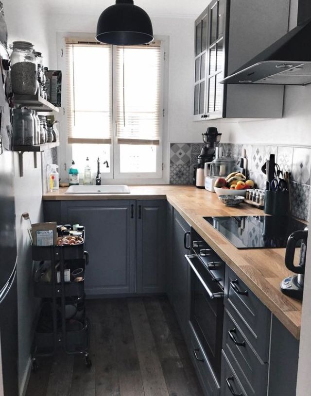 Kompaktowa kuchnia w szarościach z drewnianym blatem, oświetlona naturalnym światłem z okna. W tle widoczna półka z słoikami i wózek kuchenny.