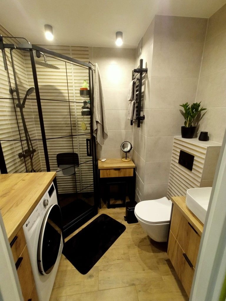 Nowoczesna łazienka z kabiną prysznicową typu walk-in, drewnianymi akcentami i czarnymi dodatkami. Widoczna pralka, umywalka nablatowa i grzejnik drabinkowy.
