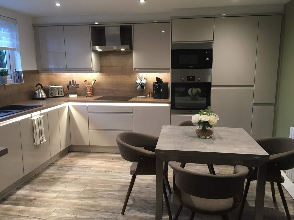 Nowoczesna kuchnia w odcieniach szarości z drewnianymi akcentami, oświetleniem LED i stołem z kwiatami. Widok na zabudowę kuchenną i strefę jadalną.