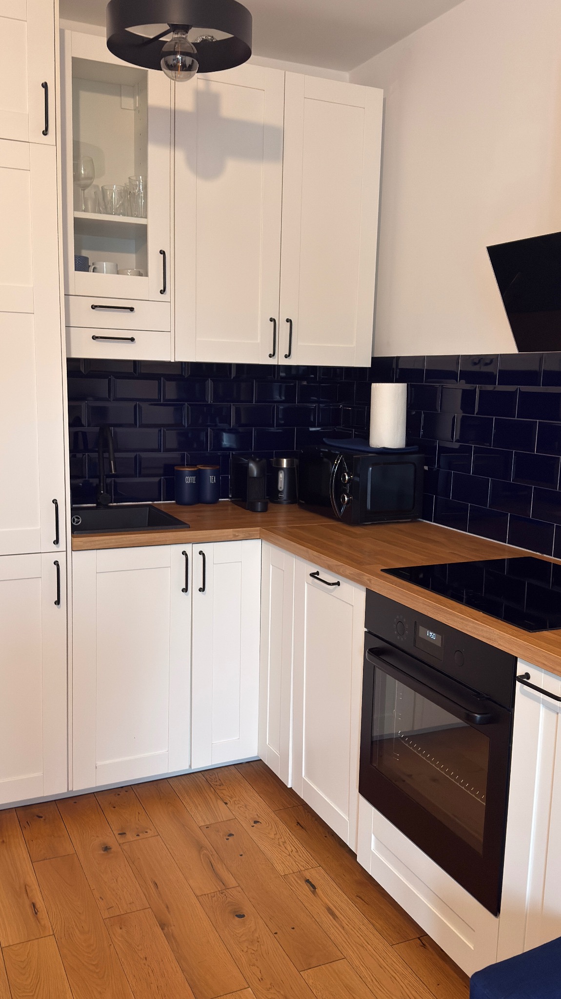 Nowoczesna kuchnia z białymi szafkami, drewnianym blatem i granatowymi płytkami. Widoczny piekarnik, mikrofalówka i akcesoria kuchenne. Parkiet z naturalnego drewna.