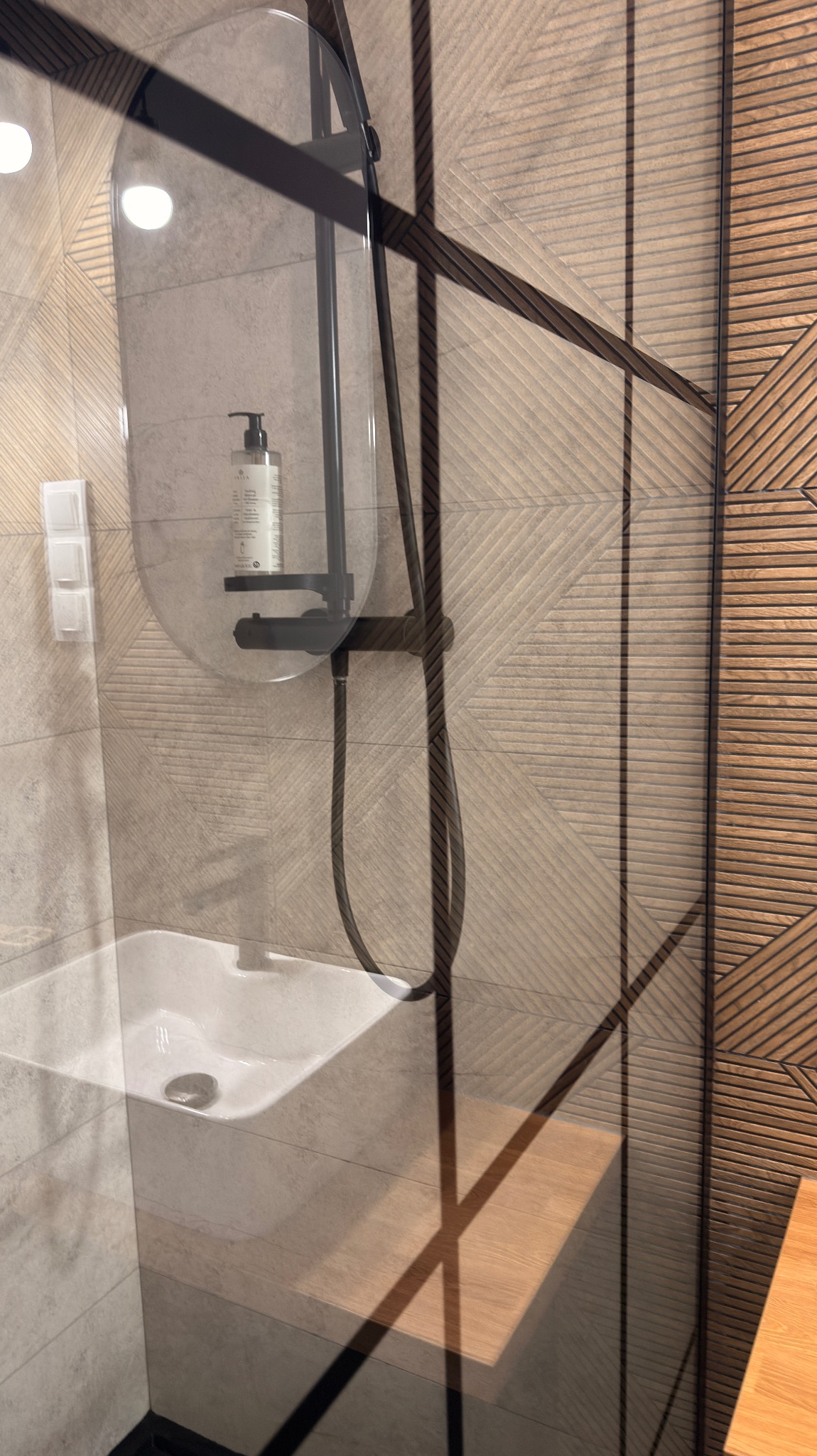 Nowoczesna kabina prysznicowa z deszczownicą i półką na kosmetyki, widoczna umywalka i drewniane panele ścienne z geometrycznym wzorem. Minimalistyczny design.