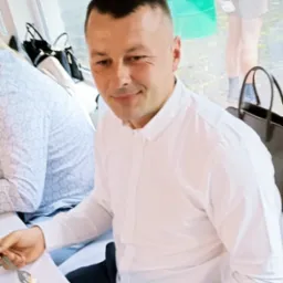 Mężczyzna w białej koszuli siedzi przy stole z resztkami ciasta na talerzu. W tle widoczne okno i zielone beczki.