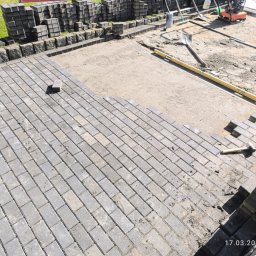 Arekbruk. - Układanie szarej kostki brukowej na podsypce piaskowej. Częściowo ułożona nawierzchnia, w tle stosy kostki i narzędzia brukarskie. Widok z góry.