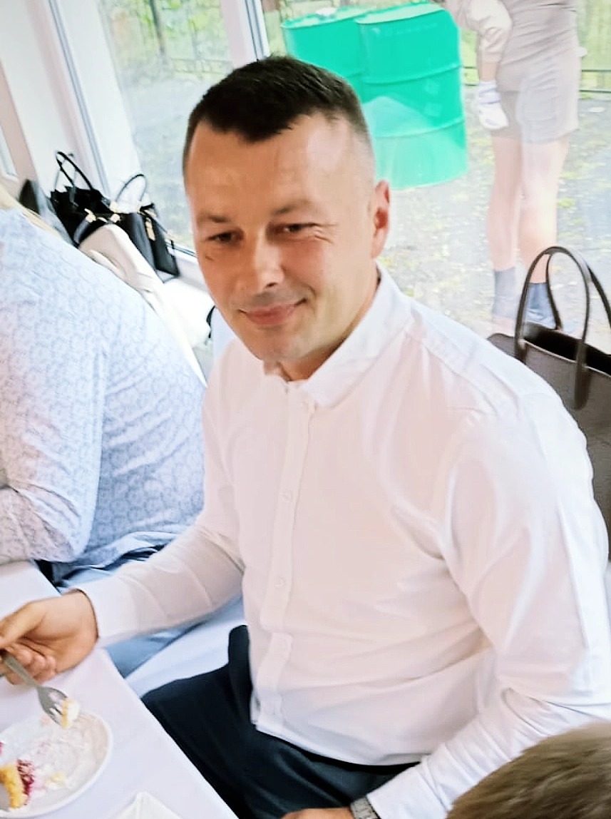 Mężczyzna w białej koszuli siedzi przy stole z resztkami ciasta na talerzu. W tle widoczne okno i zielone beczki.