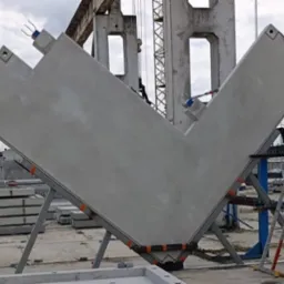 Prefabrykowany, betonowy element konstrukcyjny w kształcie litery V, wsparty na metalowej konstrukcji, z drabinką w tle. Elementy do budowy domu z keramzytu.