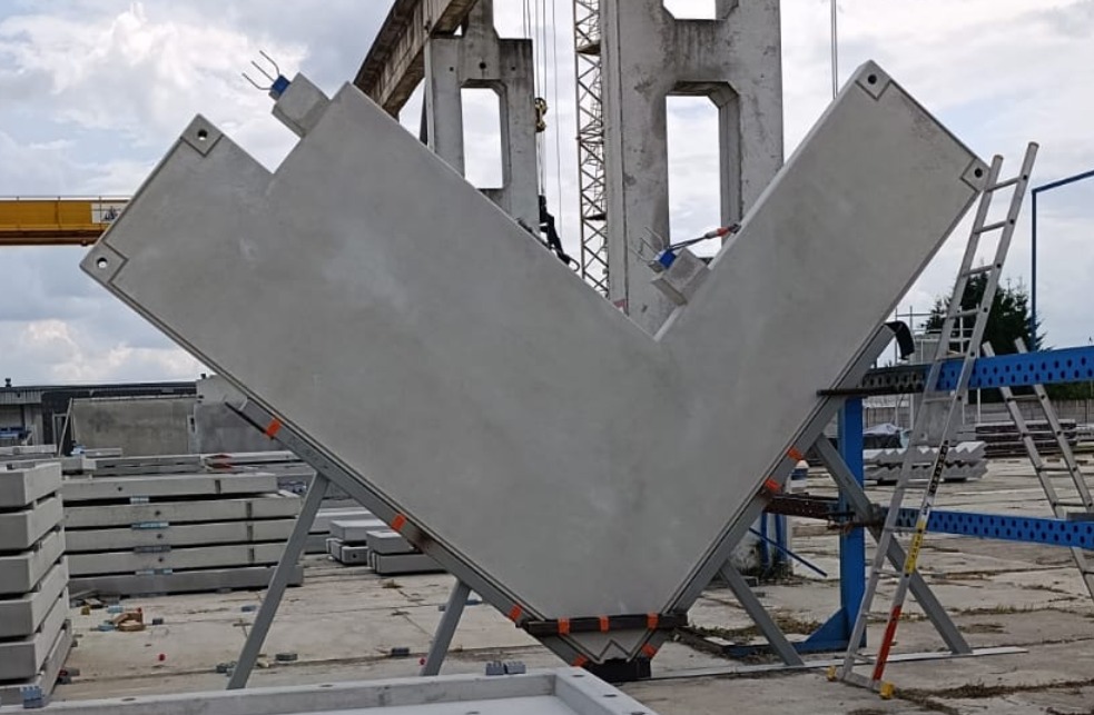 Prefabrykowany, betonowy element konstrukcyjny w kształcie litery V, wsparty na metalowej konstrukcji, z drabinką w tle. Elementy do budowy domu z keramzytu.