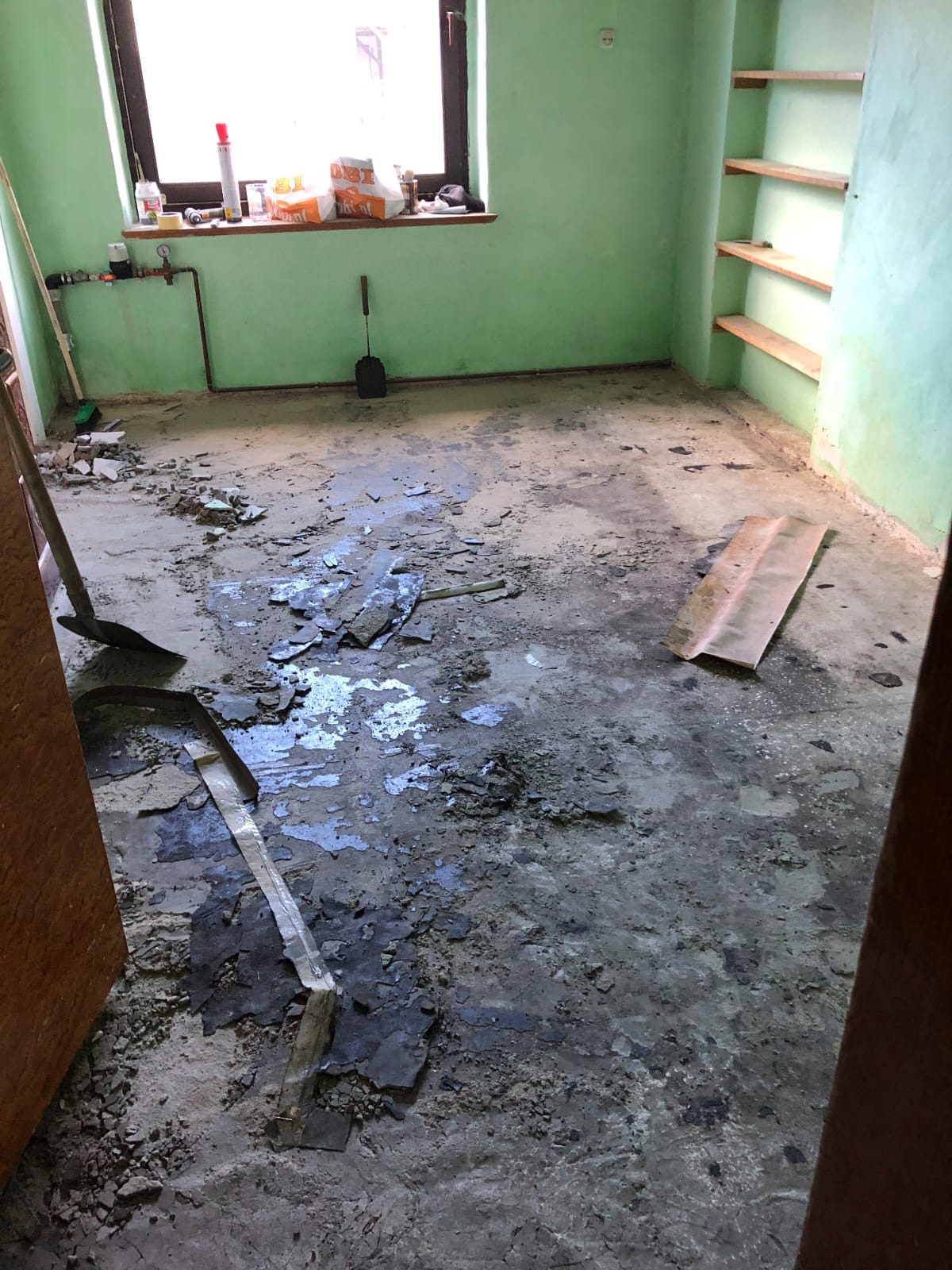 Pokój w trakcie remontu: zielone ściany, zdemontowana podłoga, widoczne resztki starej posadzki, narzędzia i materiały budowlane w tle, półki na ścianie.