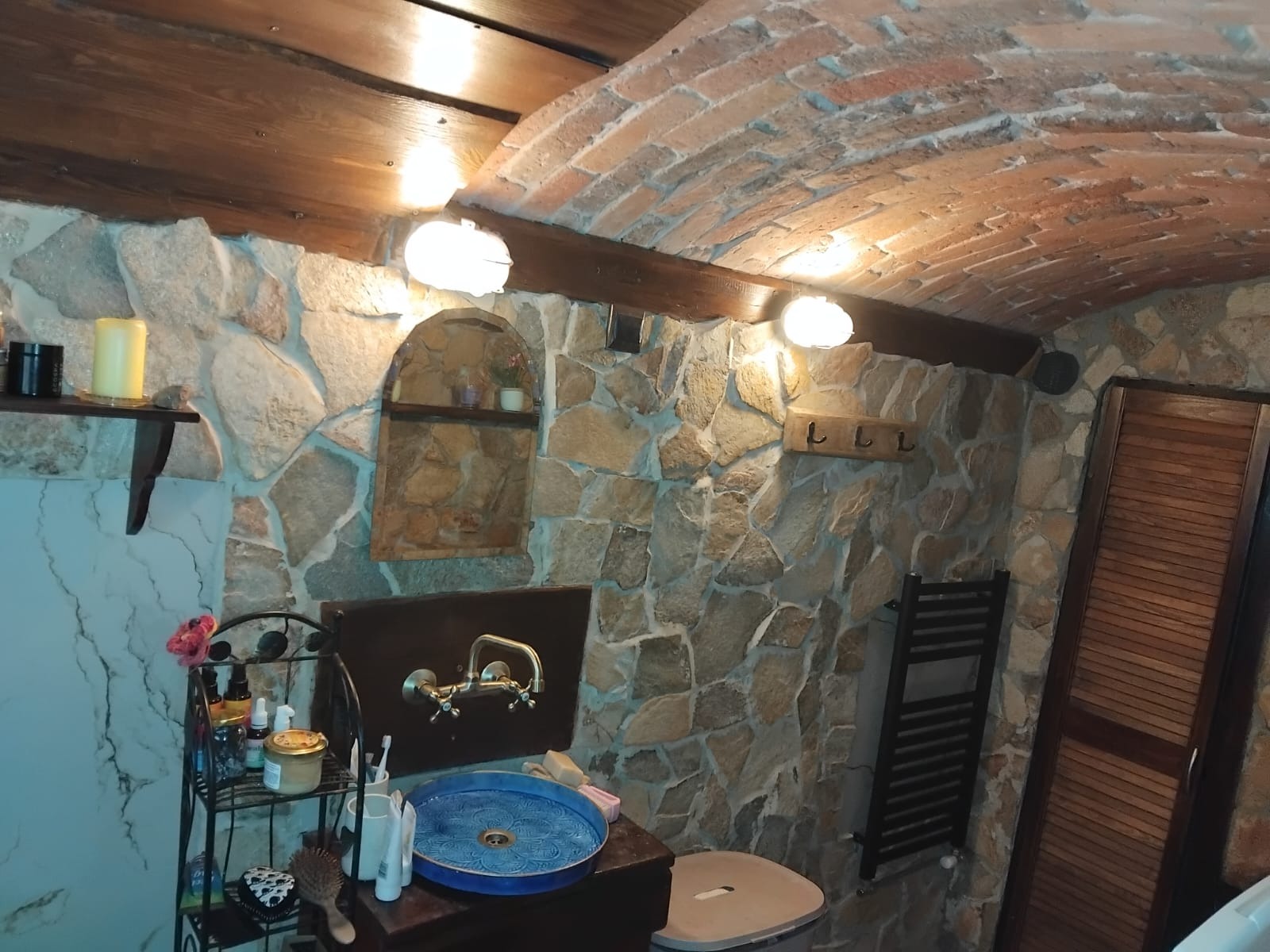 Rustykalna łazienka z kamiennymi ścianami, drewnianym sufitem z cegły i niebieską umywalką. Oświetlenie punktowe podkreśla unikalny charakter wnętrza.