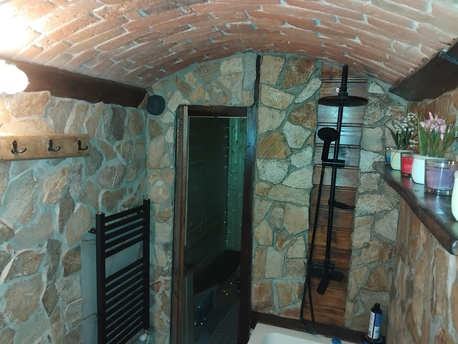 Stylowa łazienka z kamiennymi ścianami i ceglanym sklepieniem. Ciemne dodatki: grzejnik, prysznic i drewniane elementy. W tle widoczny prysznic.