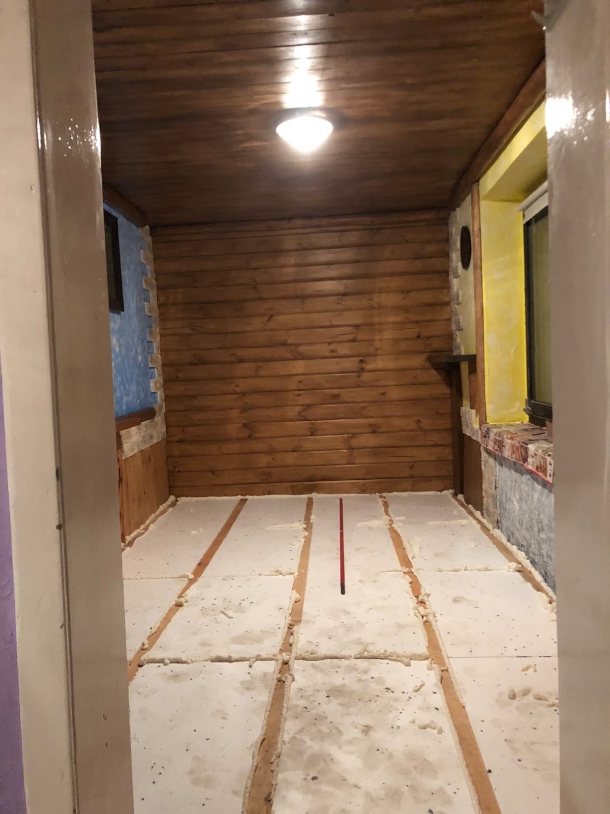 Pokój w trakcie prac wykończeniowych: drewniana boazeria, żółta ściana obok okna, podłoga z izolacją, listwy. Widok zza futryny drzwi.