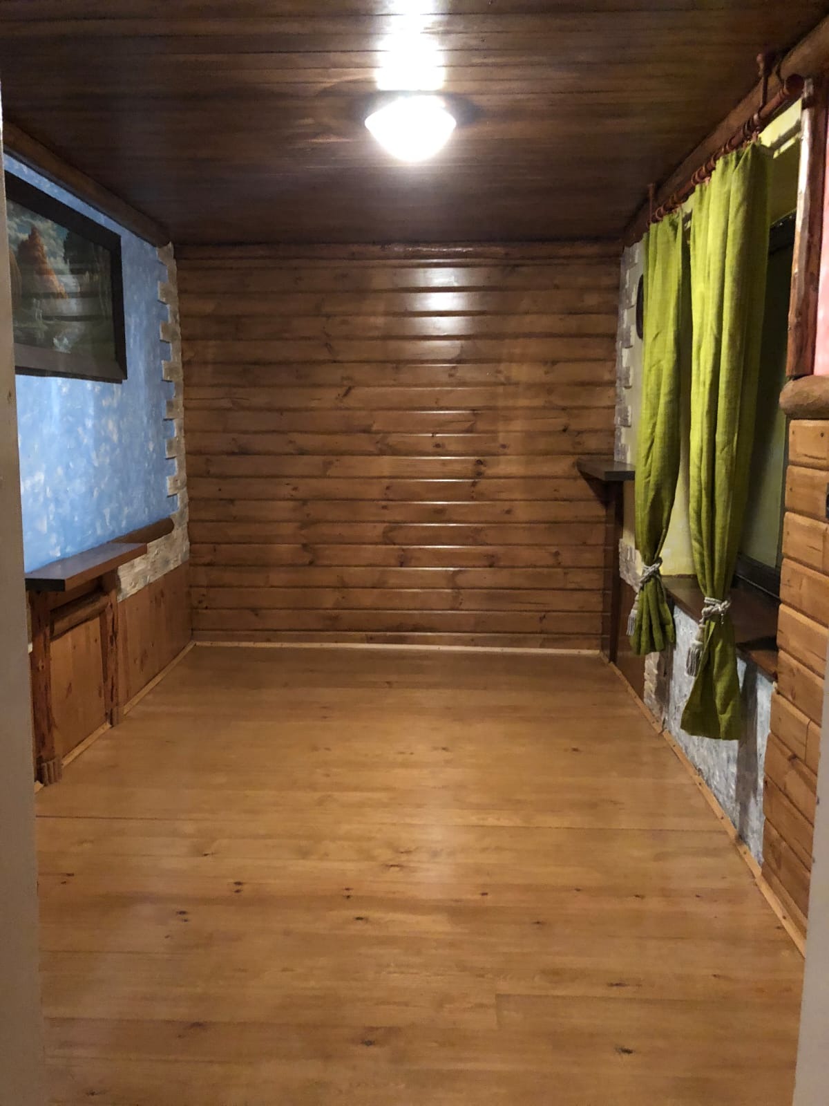 Pokój z drewnianą boazerią, jasną podłogą i elementami dekoracyjnymi z kamienia. Zielone zasłony przy oknie dodają koloru wnętrzu. Na ścianie obraz w ciemnej ramie.