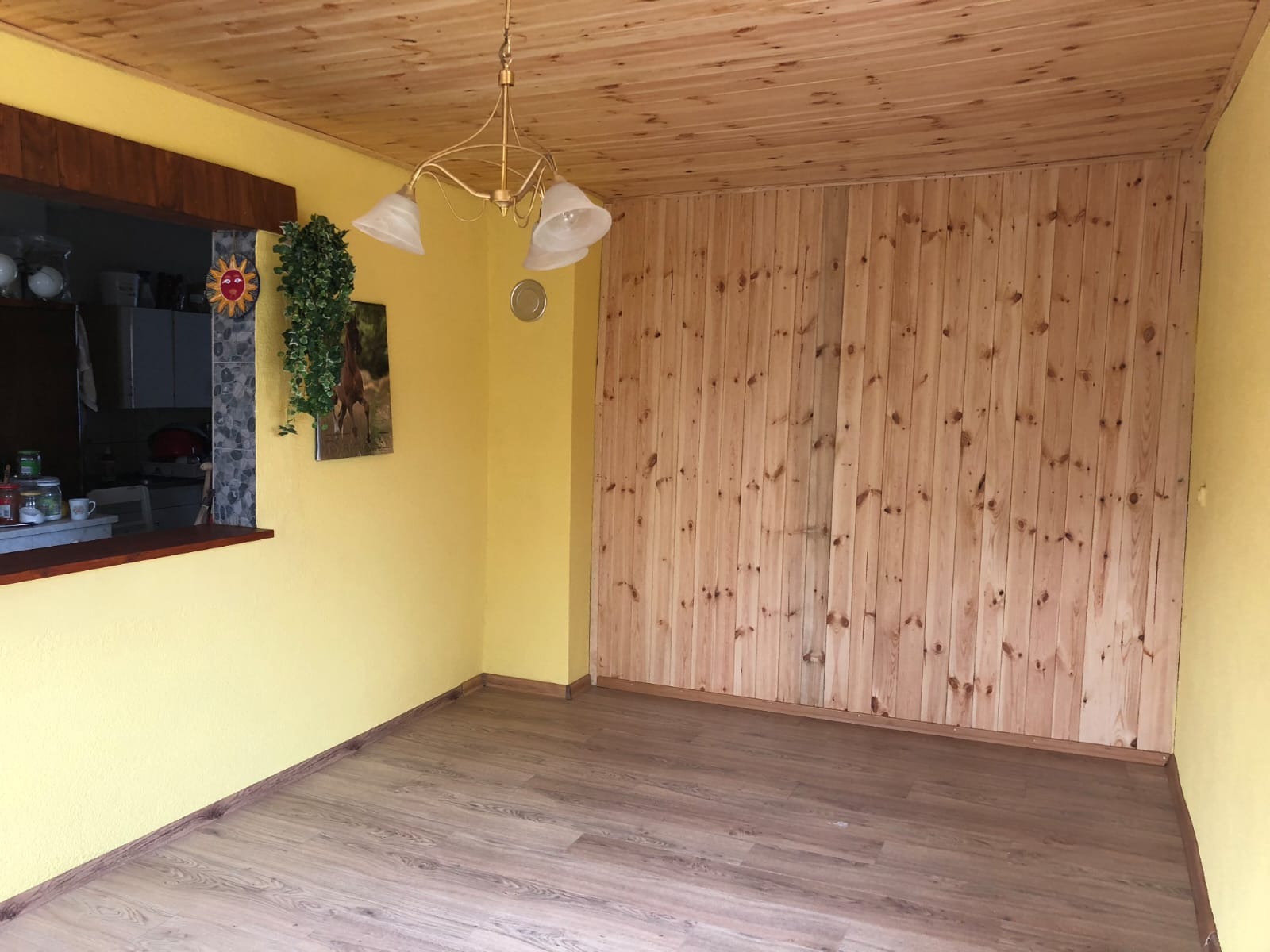 Jasne wnętrze z boazerią, żółtymi ścianami i drewnianą podłogą. Widoczne okno z dekoracjami i lampa wisząca. Świeżo wykończone pomieszczenie.