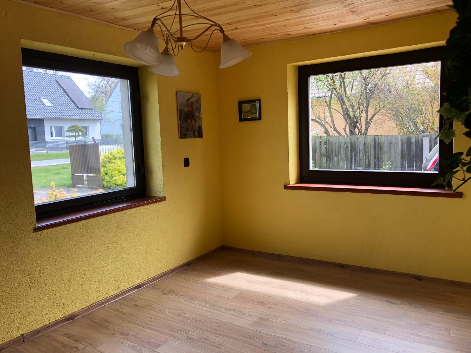 Żółty pokój po malowaniu z drewnianą podłogą i dwoma oknami z widokiem na zewnątrz, oświetlony przez wiszącą lampę. Dekoracje na ścianach.