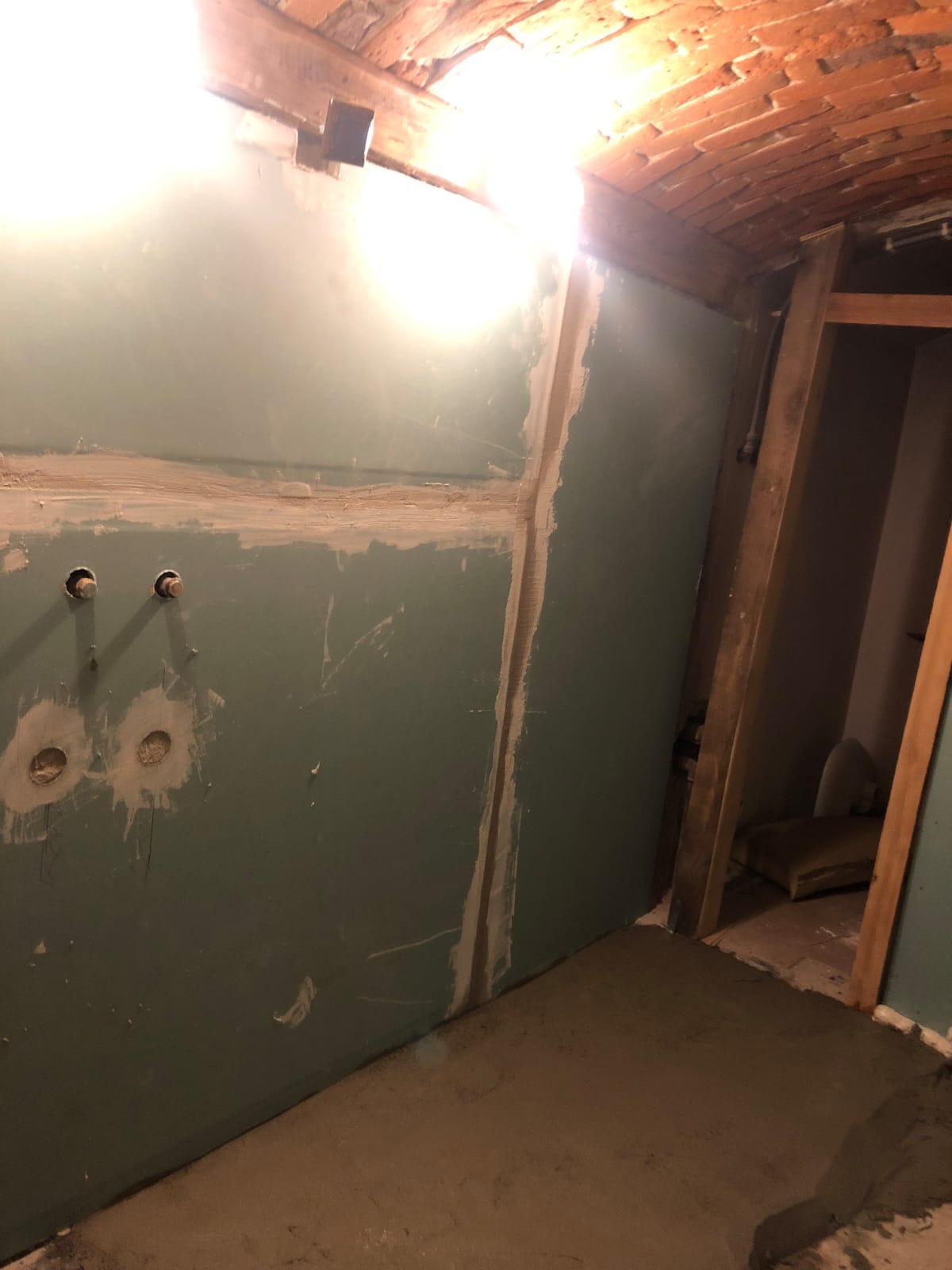 Narożnik pomieszczenia w trakcie remontu: ściany z płyt kartonowo-gipsowych, częściowo zaszpachlowane, surowa cegła na sklepieniu i wylewka podłogowa.