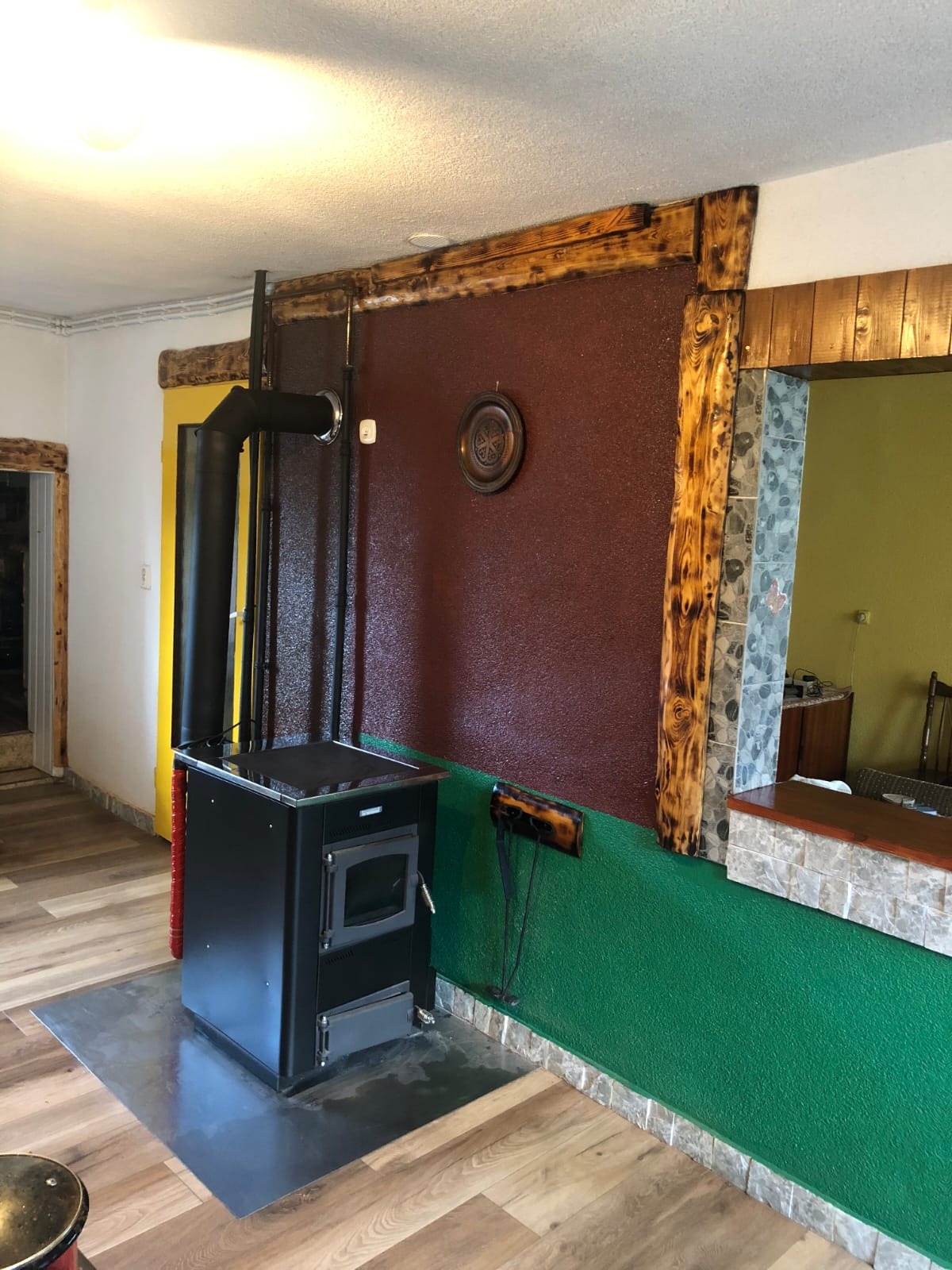 Rustykalny piec wolnostojący na metalowej płycie, z czarną rurą odprowadzającą dym. Ściany w odcieniach brązu i zieleni, drewniane wykończenia.