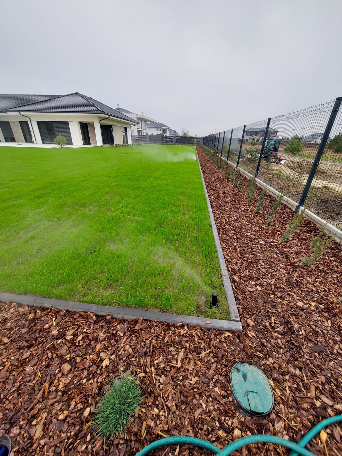 Automatyczne nawadnianie trawnika zraszaczami w ogrodzie z korą i ogrodzeniem panelowym. W tle nowoczesny dom i traktor.