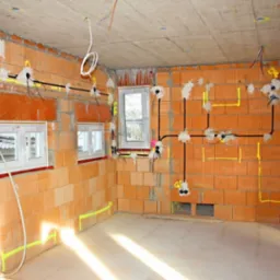 Wnętrze w trakcie prac elektrycznych: kable i puszki instalacyjne zamontowane na ceglanych ścianach, widoczne okna i betonowy sufit. Żółte oznaczenia punktów elektrycznych.