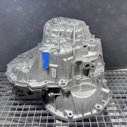 Szymon Koblowski - Szara, metalowa obudowa skrzyni biegów z niebieską taśmą, umieszczona na metalowej kratownicy na tle szarej ściany. Widoczne otwory montażowe i numery seryjne.