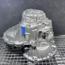 Szara, metalowa obudowa skrzyni biegów z niebieską taśmą, umieszczona na metalowej kratownicy na tle szarej ściany. Widoczne otwory montażowe i numery seryjne.