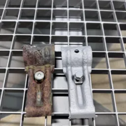 Porównanie uchwytów: zardzewiały i po piaskowaniu, leżące na metalowej kracie. Widoczna różnica w wyglądzie i stanie technicznym elementów.