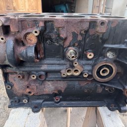 Szymon Koblowski - Blok silnika spalinowego, czarny z rdzawymi nalotami, umieszczony na drewnianych podporach. Widoczne otwory i detale konstrukcyjne.