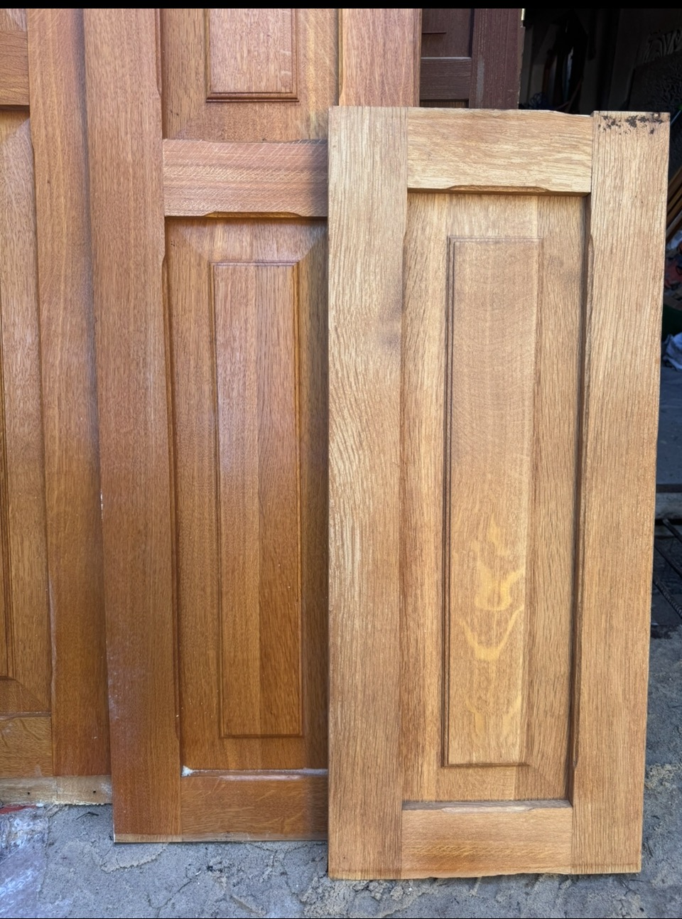 Drewniane, fornirowane fronty meblowe w odcieniach brązu, z widoczną strukturą drewna i subtelnymi zdobieniami, ułożone na szarym podłożu. Elementy czekają na montaż.