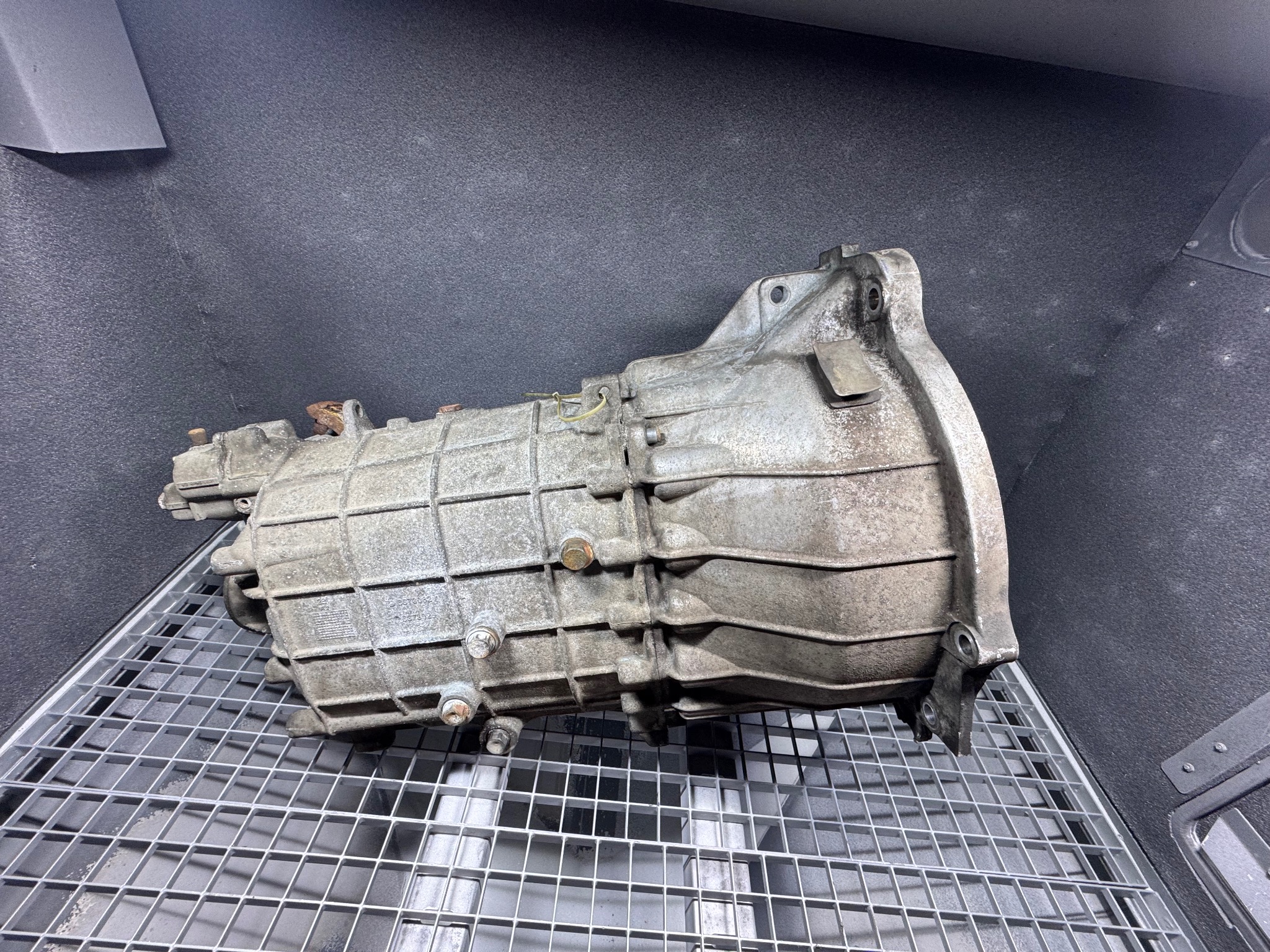 Szara, używana skrzynia biegów na metalowej kratce, z widocznymi śrubami i elementami konstrukcyjnymi. Tło stanowi ciemna, teksturowana powierzchnia.