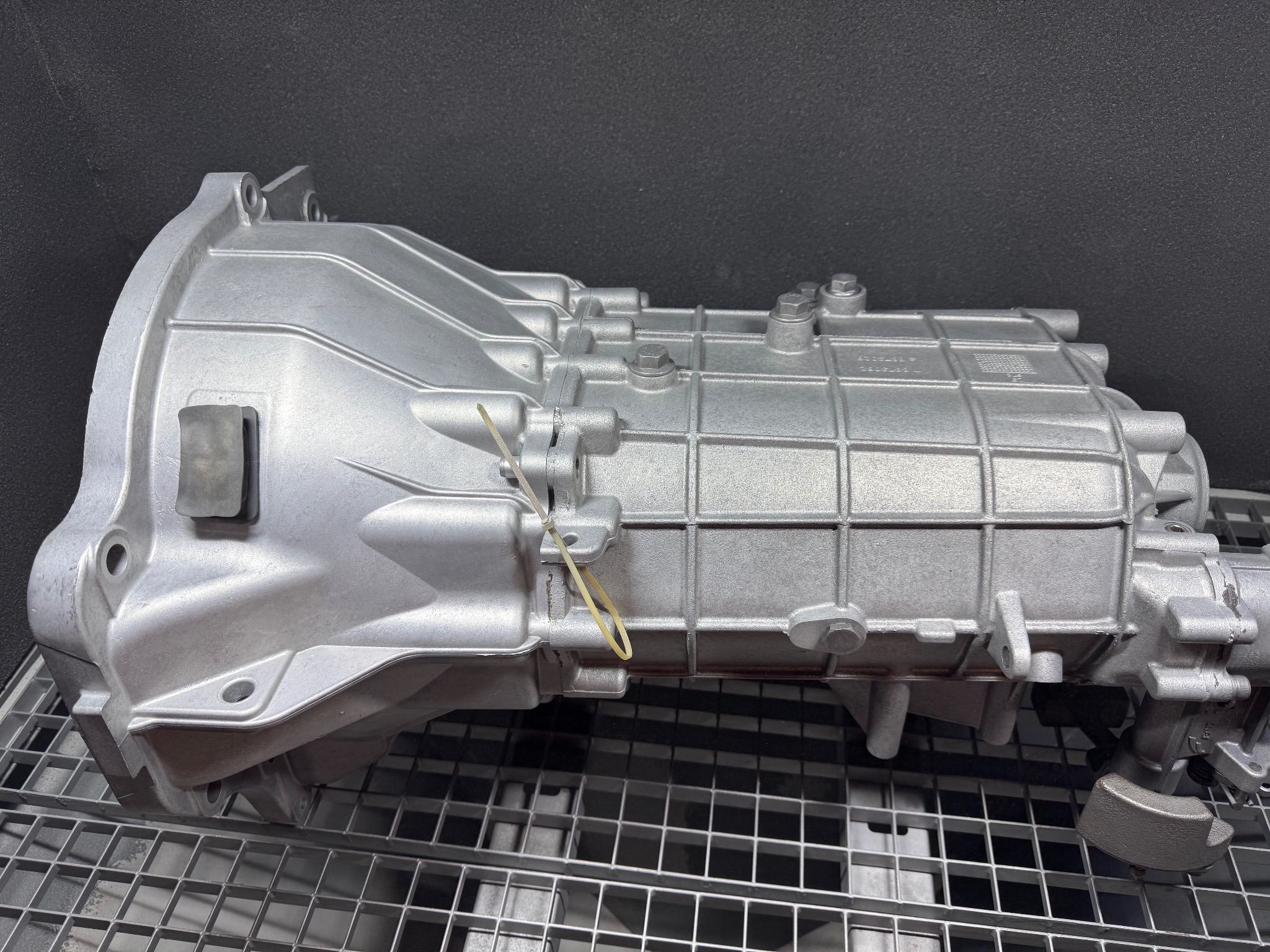 Szara, metalowa skrzynia biegów na metalowej kratce, z widocznymi śrubami i elementami konstrukcyjnymi, oświetlona studyjnie, na ciemnym tle.