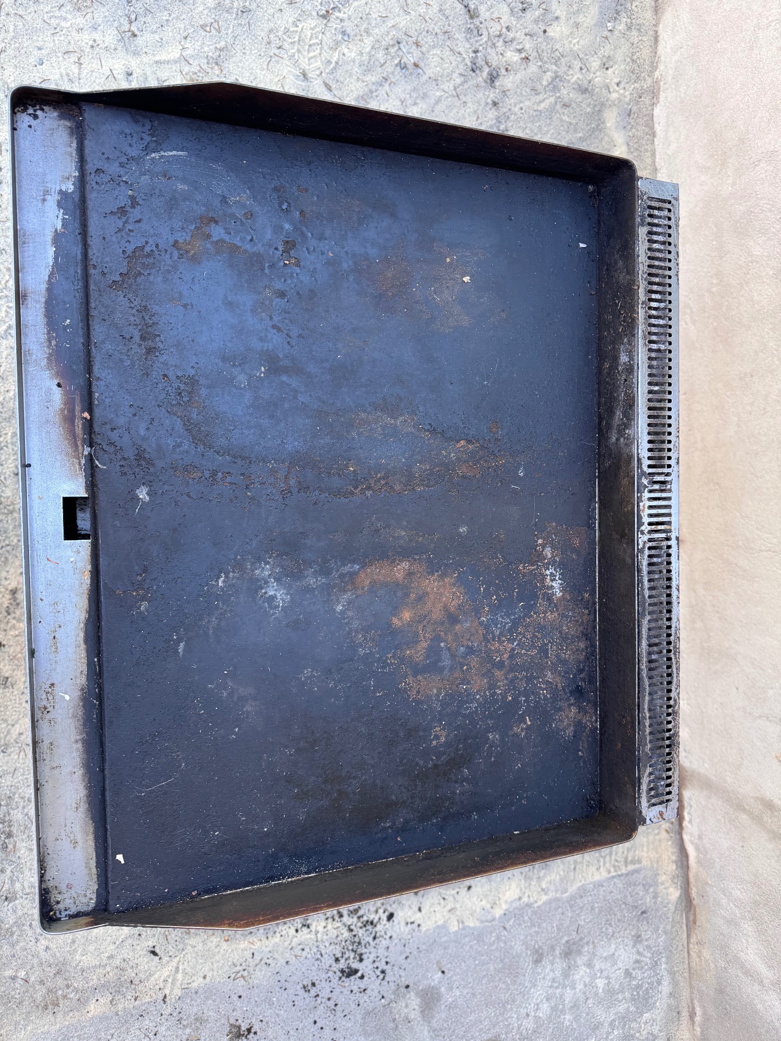 Brudna, prostokątna blacha do pieczenia z rusztem ociekowym po prawej stronie, leżąca na betonowym podłożu. Widoczne ślady użytkowania i przypalenia.