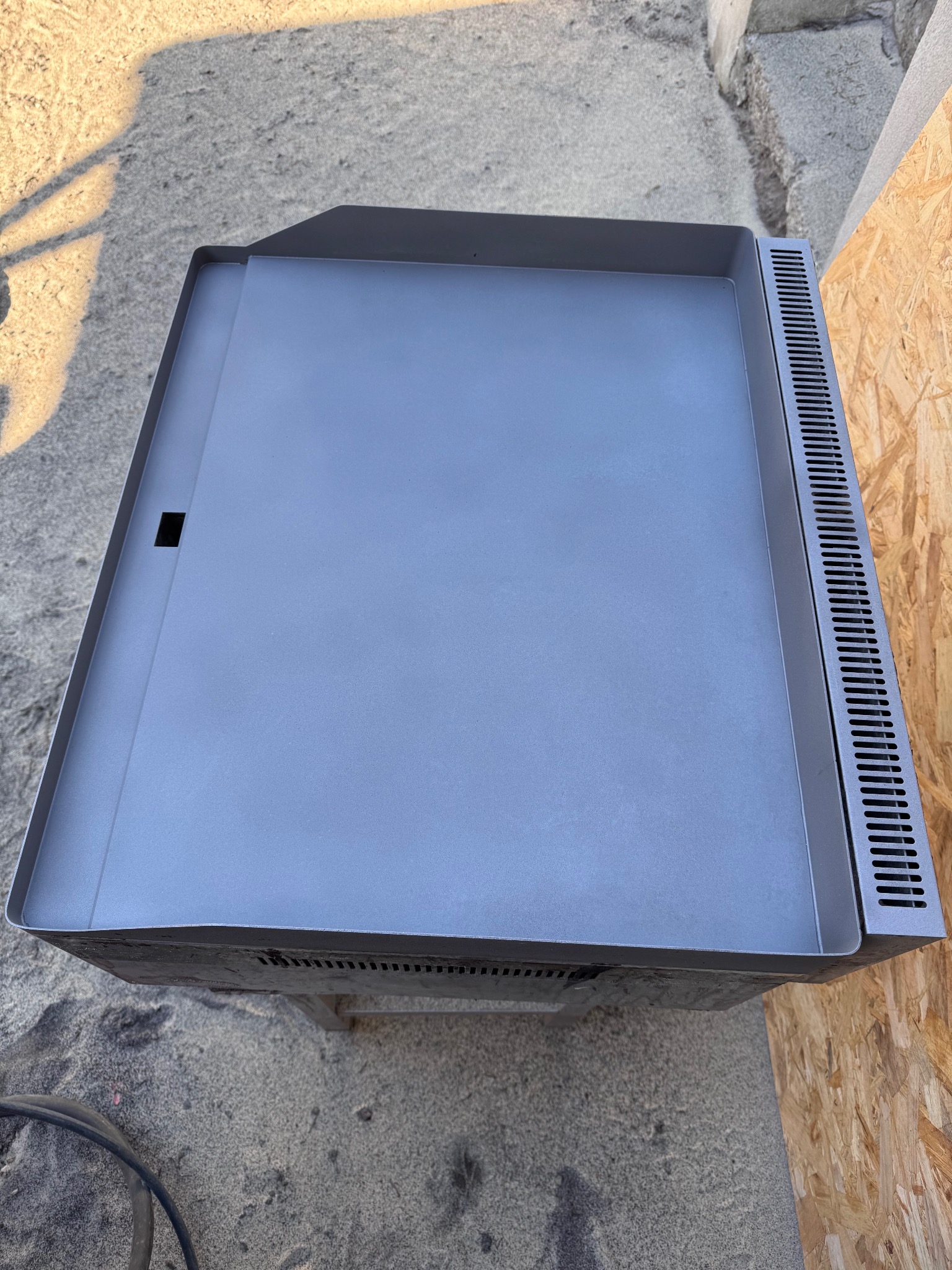 Szara, metalowa płyta grzewcza z perforowaną krawędzią, ustawiona na zewnątrz przy betonowej ścianie i płycie OSB. Widoczne ślady użytkowania.