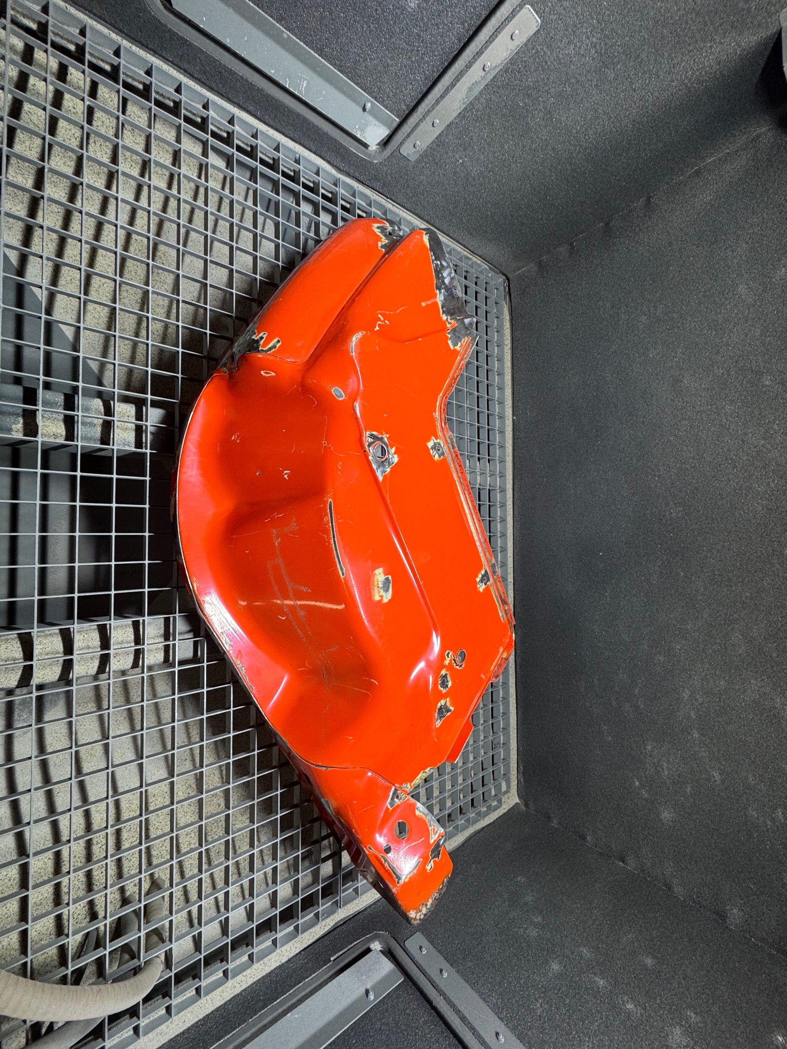 Czerwony element karoserii z uszkodzeniami w komorze piaskarki na metalowej kracie. Widoczne ślady korozji i odpryski farby. Przygotowanie do piaskowania.