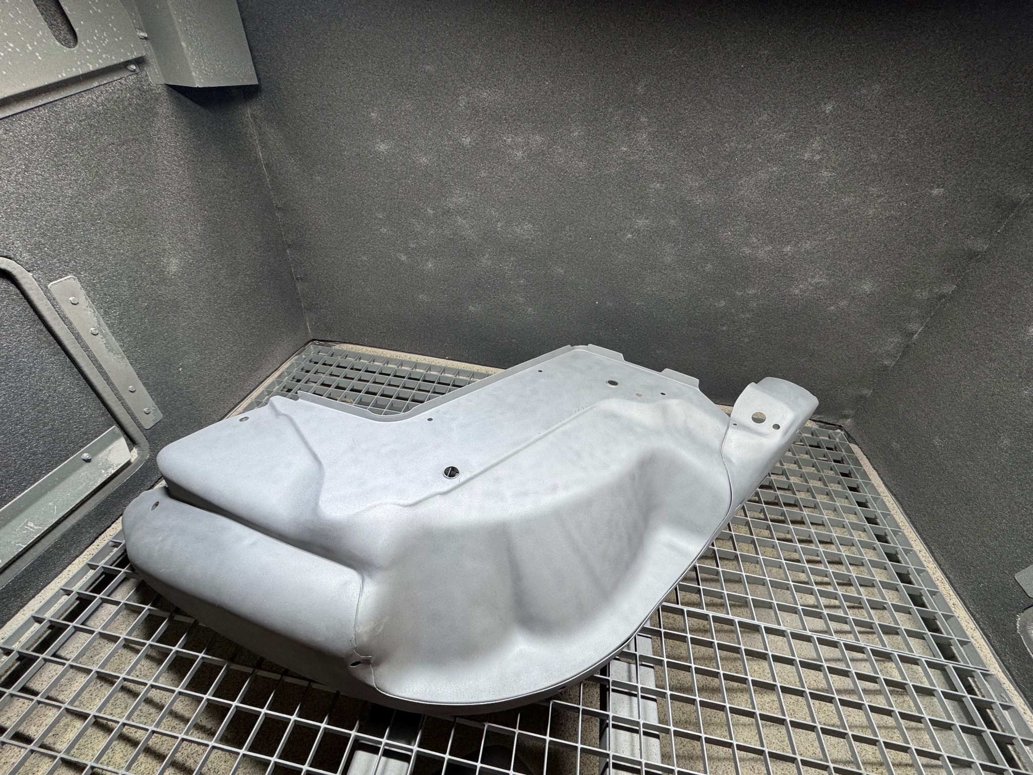 Szara, metalowa obudowa w komorze piaskarskiej, leżąca na kratce. Widoczne ślady piaskowania na ścianach komory.