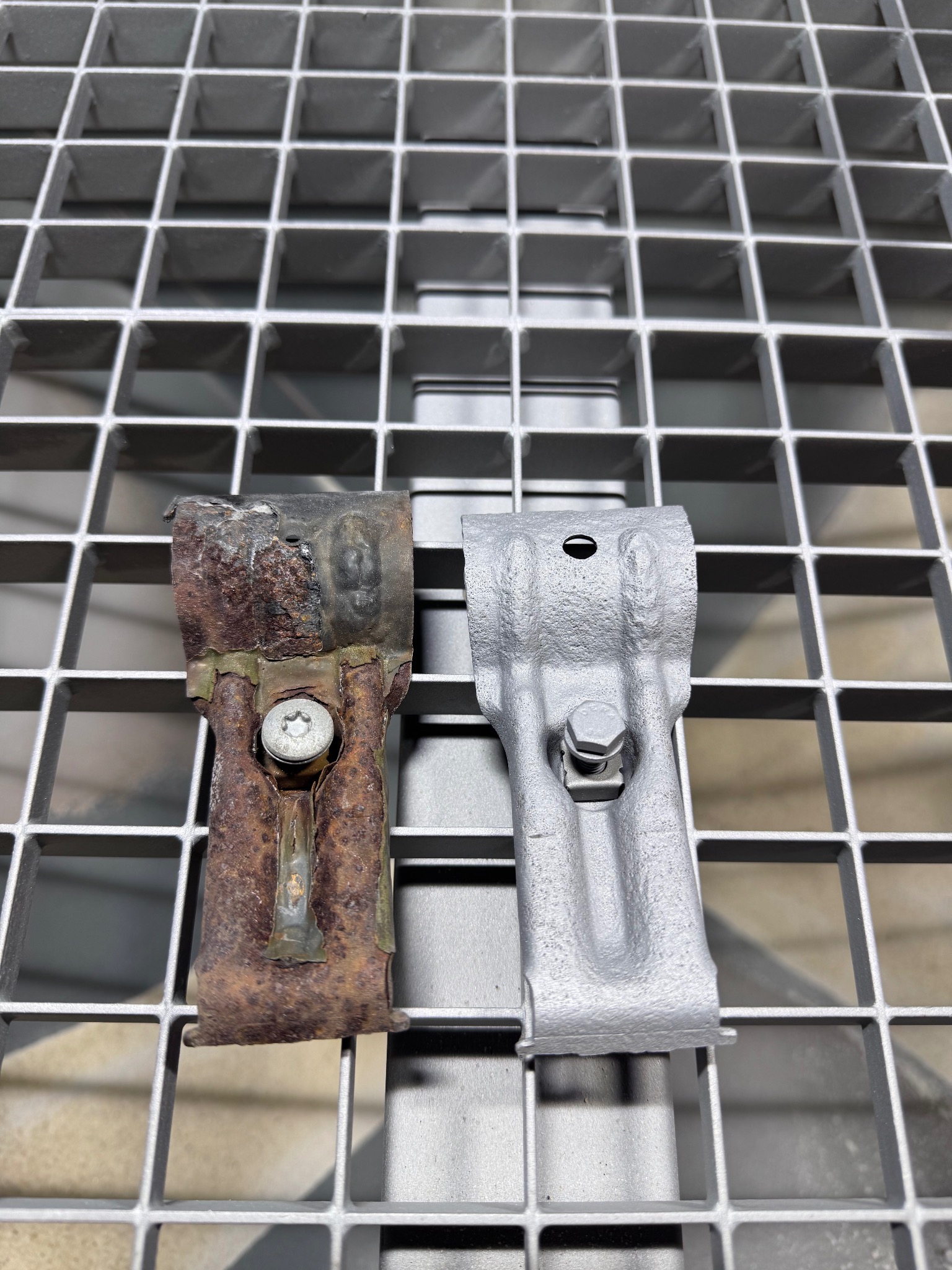 Porównanie uchwytów: zardzewiały i po piaskowaniu, leżące na metalowej kracie. Widoczna różnica w wyglądzie i stanie technicznym elementów.