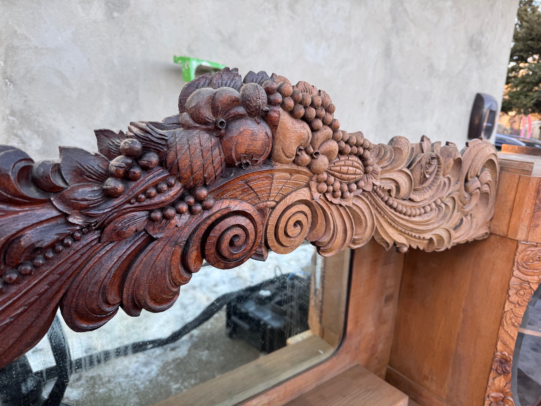 Ręcznie rzeźbione, drewniane zdobienie z motywami roślinnymi i owocowymi, prawdopodobnie fragment ramy lustra lub mebla, na tle szarej ściany.