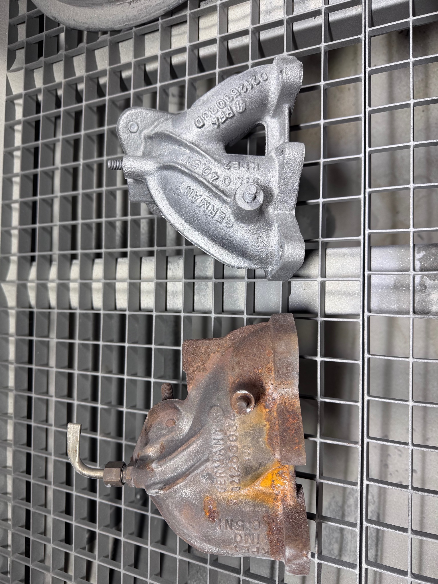 Dwie metalowe części na kratce: górna po piaskowaniu, srebrna i czysta; dolna zardzewiała, przed czyszczeniem. Widoczne detale i napisy na elementach.