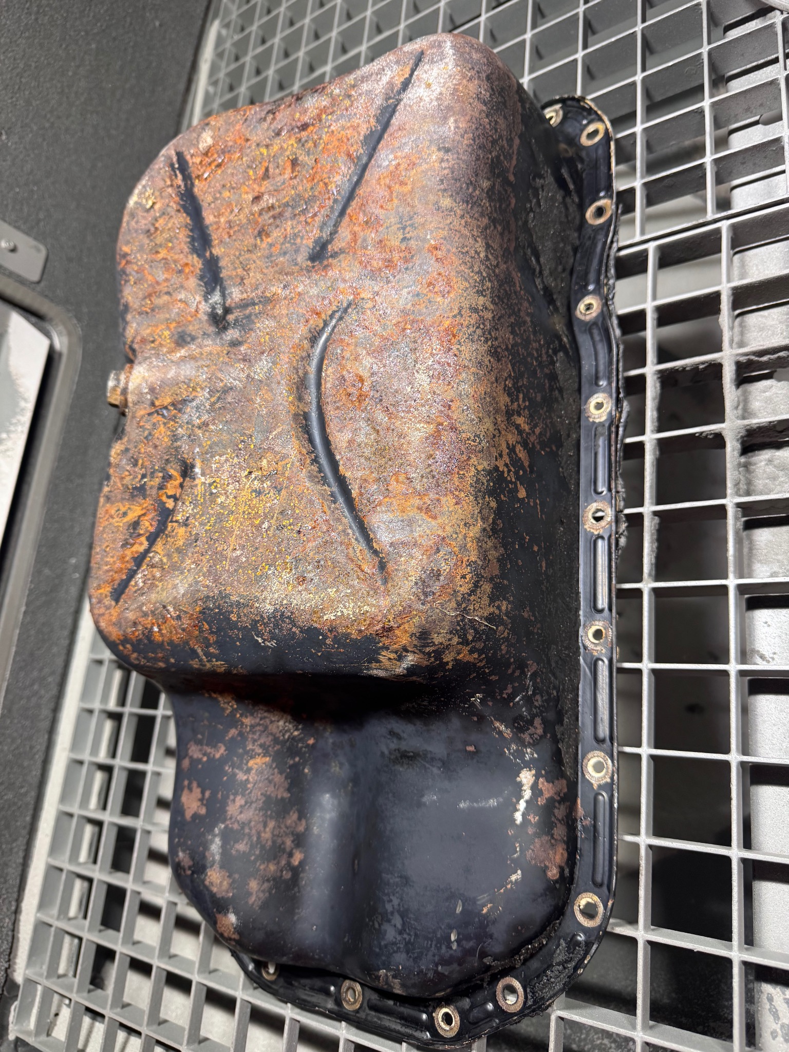 Zardzewiała, metalowa miska olejowa z widocznymi śladami korozji, leżąca na metalowej kratce. Widoczne otwory montażowe i ślady oleju.