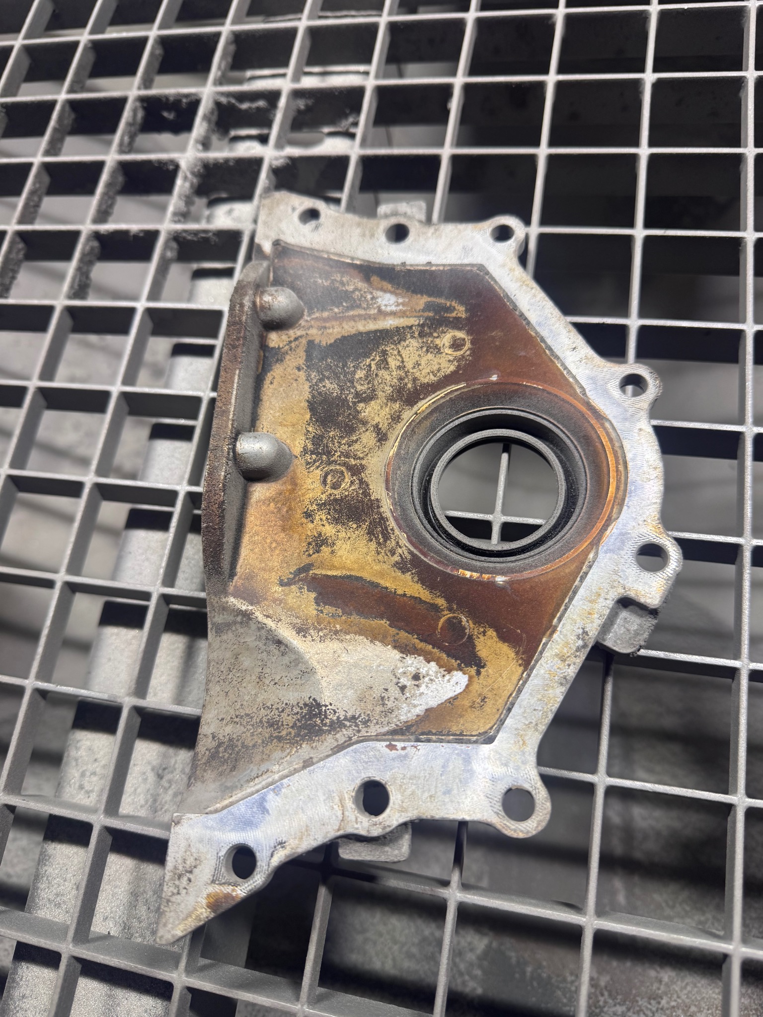 Oczyszczona, metalowa część silnika z widocznymi śladami korozji, leżąca na metalowej kratce. Widoczne otwory montażowe i okrągły otwór centralny.