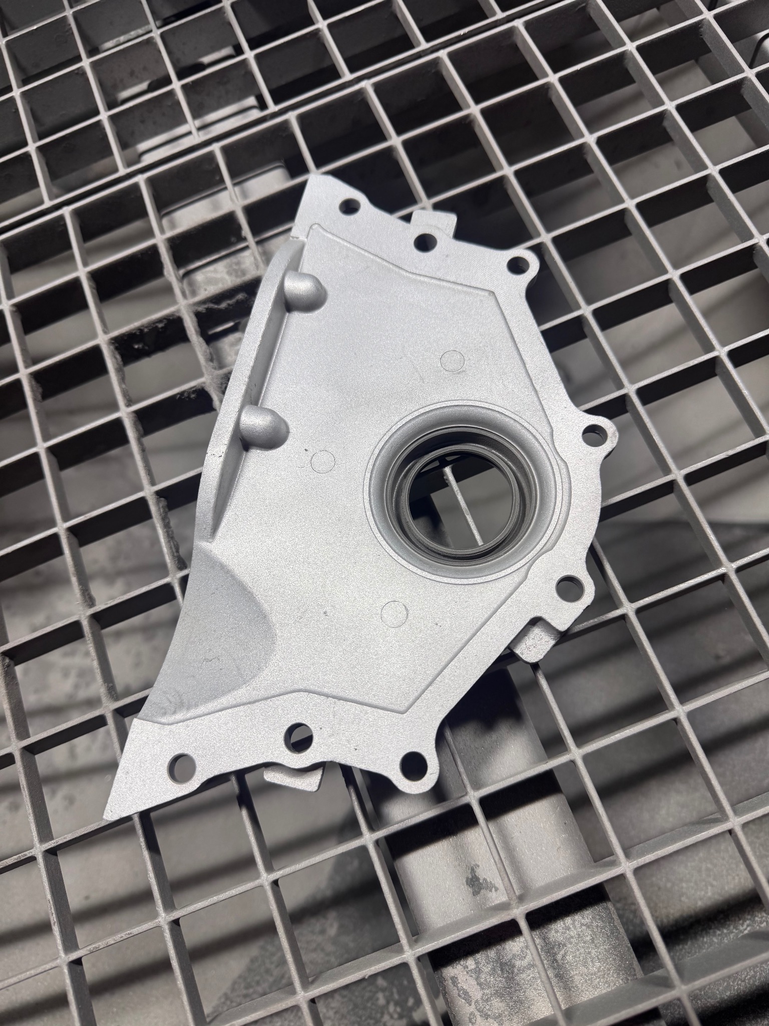 Szara, metalowa część samochodowa z otworem, leżąca na metalowej kratce. Widoczne ślady obróbki, otwory montażowe i charakterystyczny kształt elementu.