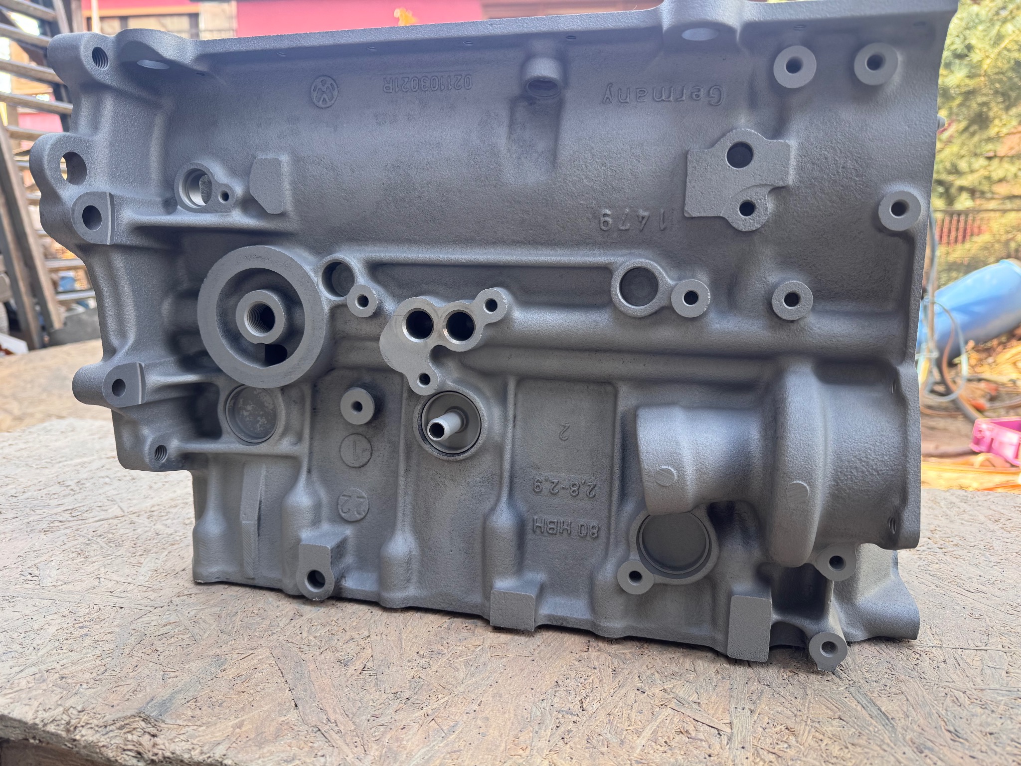 Szary, odnowiony blok silnika spalinowego z widocznymi numerami seryjnymi i oznaczeniami producenta, leżący na drewnianej powierzchni. Widoczne ślady obróbki.