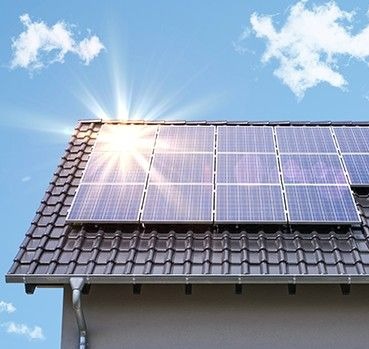 Panele słoneczne na dachu domu z ciemnej dachówki, w słoneczny dzień. Widoczny fragment rynny i elewacji. Błękitne niebo z białymi chmurami.